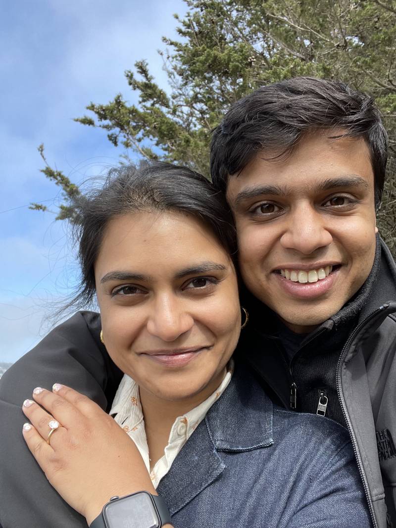 The happy couple pose in a vertical seflie, smiling at the camera, with Adhiti brandishing her engagement ring on her hand for the camera.