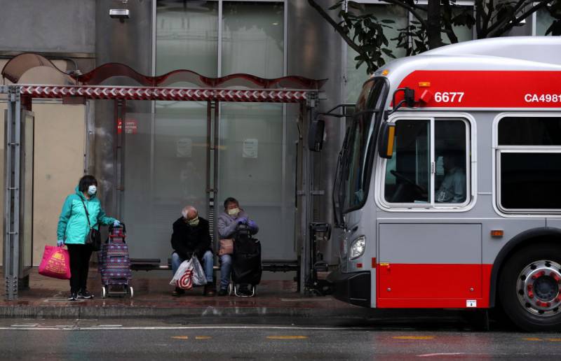 People wear masks as they wait in a bus shelter as a red and gray bus pulls up.