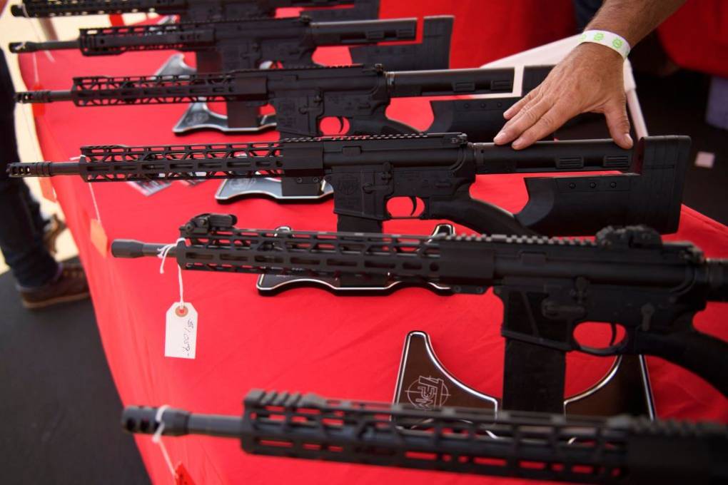 Assault weapons on display on a red tablecloth as a hand rests on top of one weapon.
