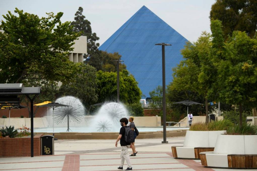 A young man is walking across a campus with a blue pyramid building, fountain and trees in the background.