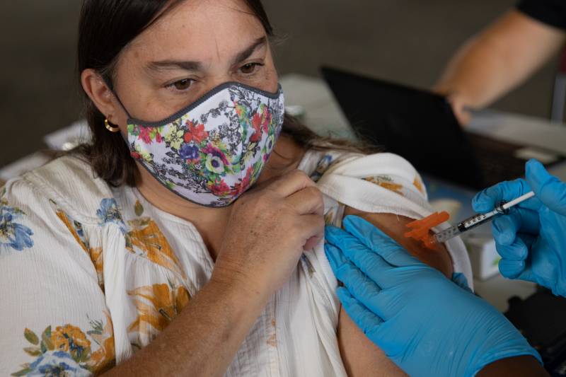 A woman wearing a floral mask over her mouth and nose and a floral shirt holds up her left sleeve as someone wearing blue gloves injects her upper arm with a needle.