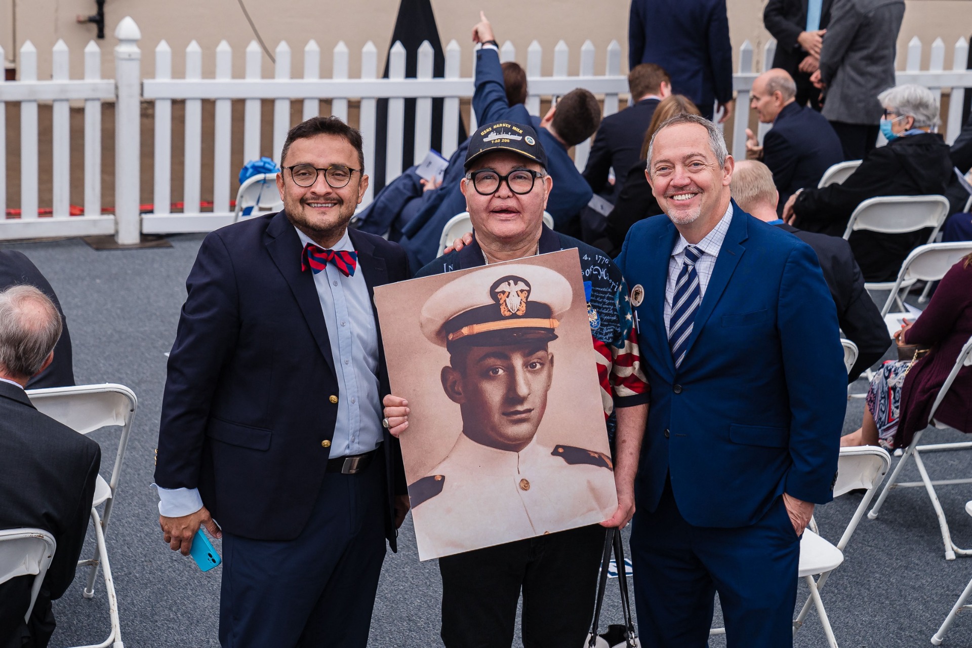 Three men stand together outside at an event with white chairs in the background. From left to right, a man in a bow tie and suit, another man in a navy hat and glasses holding a photo of Harvey Milk in naval uniform, and right, a smiling man in a suit with a goatee.