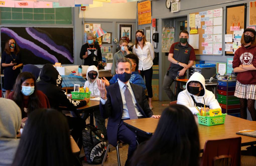 A man wearing a mask and business suit sits in a classroom at a desk surrounded by young adults wearing masks.
