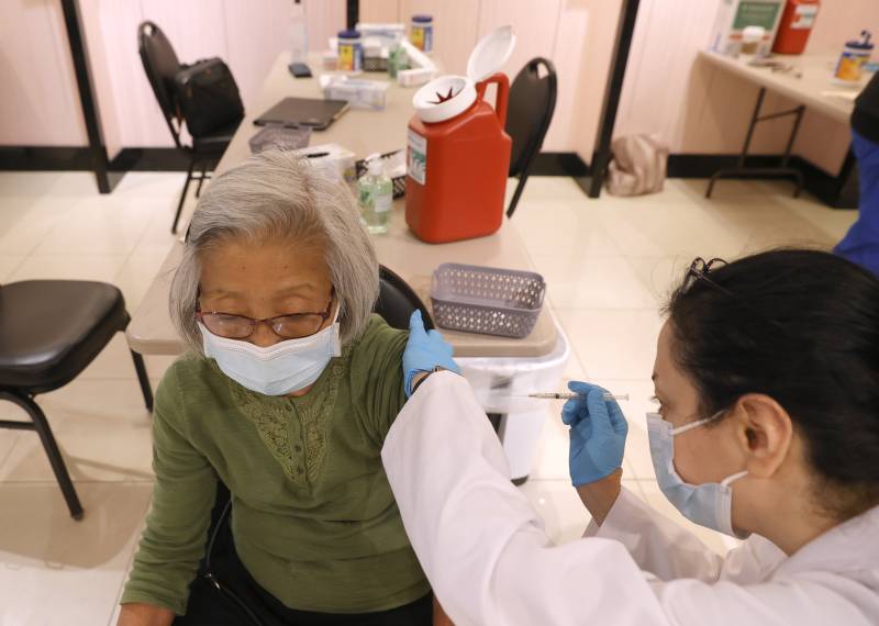 A woman in a mask receives a shot from a another woman in a white lab coat.