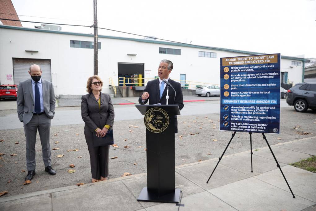 California Attorney General Rob Bonta stands at a lectern in front of a large warehouse, flanked by two people on his right and a COVID safety sign on his left.