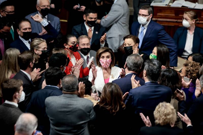 Nancy Pelosi stands, in a white jacket, amid a crowd of lawmakers.