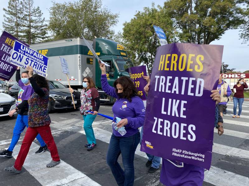 People wearing purple cross a street carrying protest signs, one reads, "heroes treated like zeroes."
