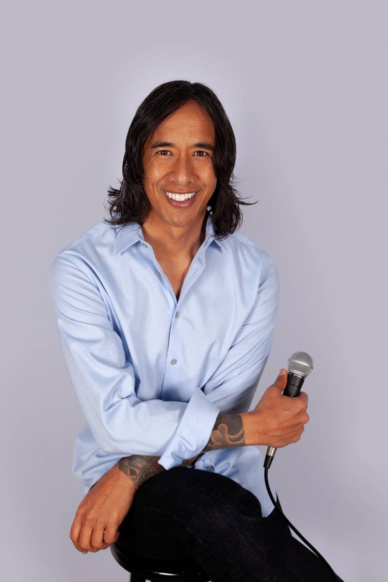 A man who describes himself as mixed race poses for a studio-style photo against a light gray background. He's got shoulder-length black hair and is smiling wide, in a blue collared shirt with his arms crossed, holding a microphone. He has "sleeve" tattoos visible because his shirt cuffs are rolled up just a bit. He holds a microphone in one hand.