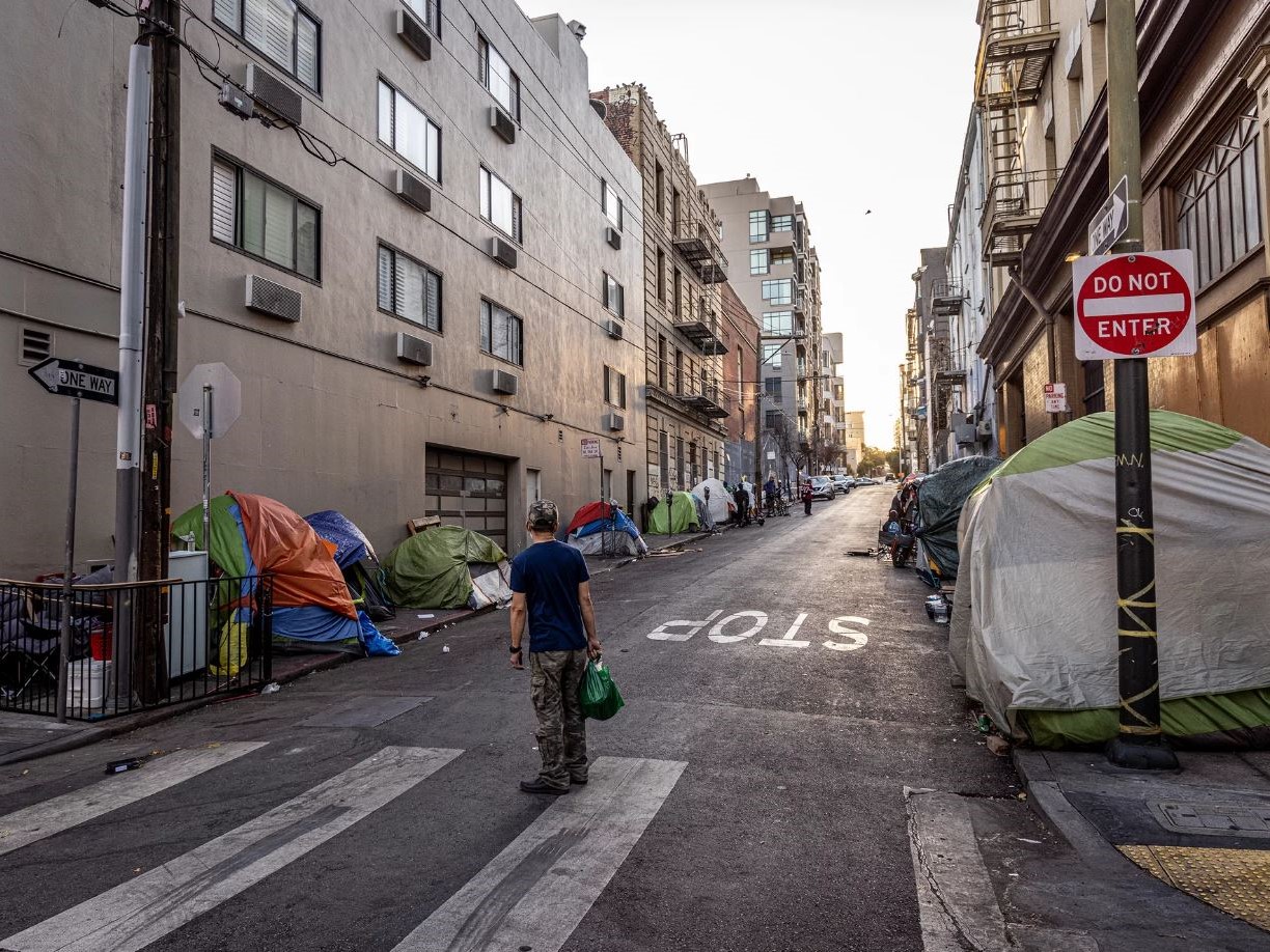 A person stands in the middle of a crosswalk looking up the street and at a row of tents on each sidewalk.