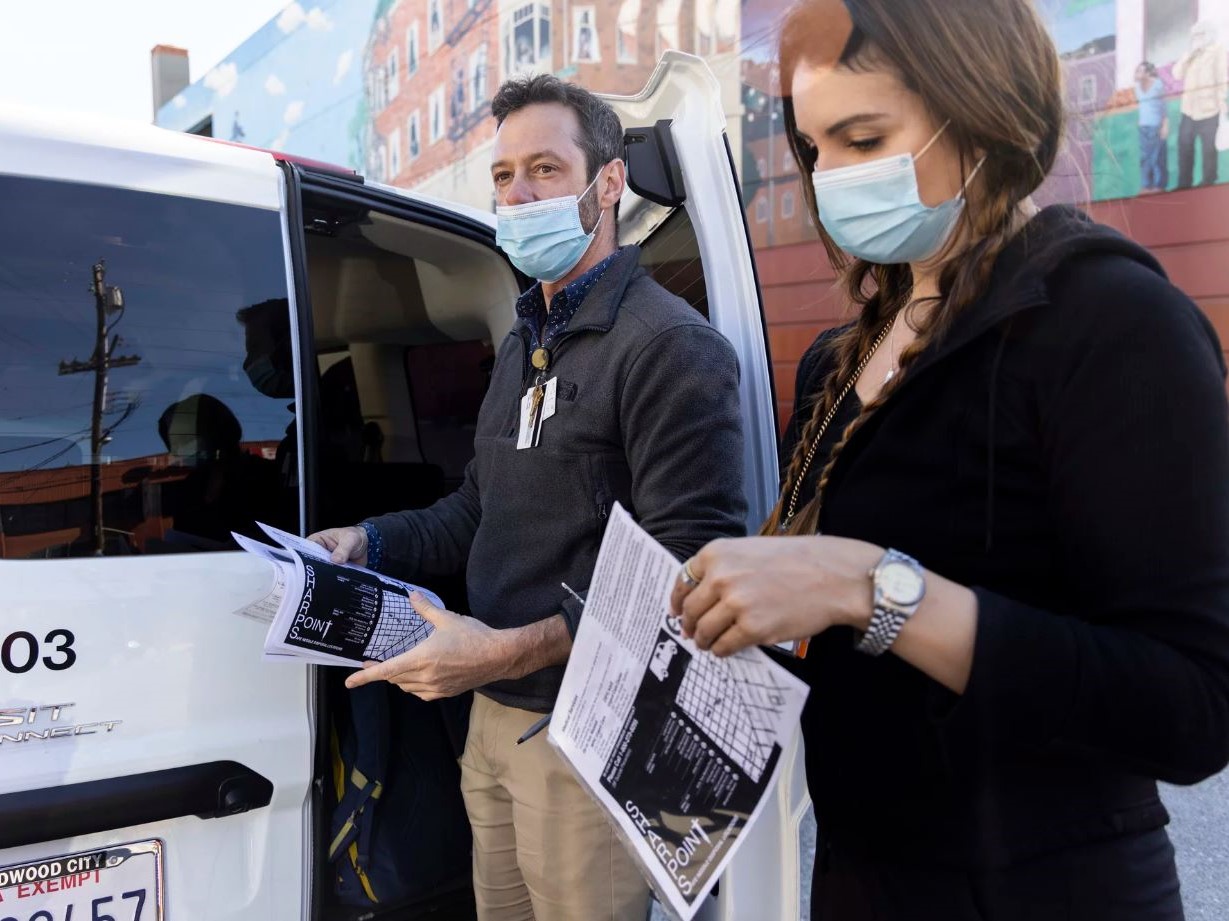 Two people wearing face masks stand outside a white van parked alongside a mural as they look through papers and plastic bags in their hands. It looks like early morning.