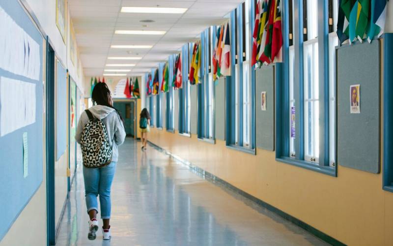 A high school student with a backpack walks down a long, windowed school hallway.