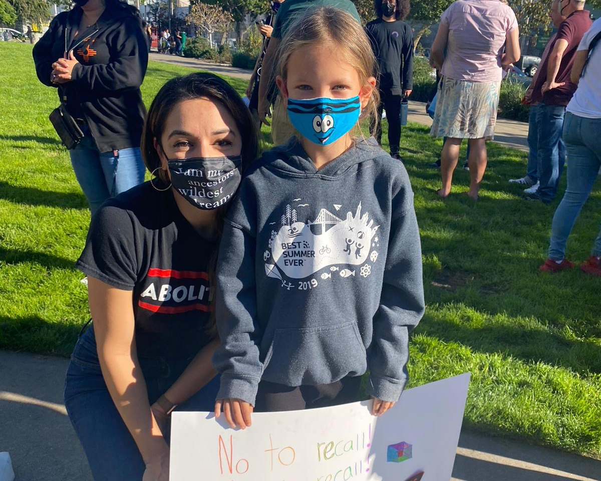 A woman wearing a black mask kneels down next to a small girl wearing a blue mask who is holding a poster that reads "no to recall!"