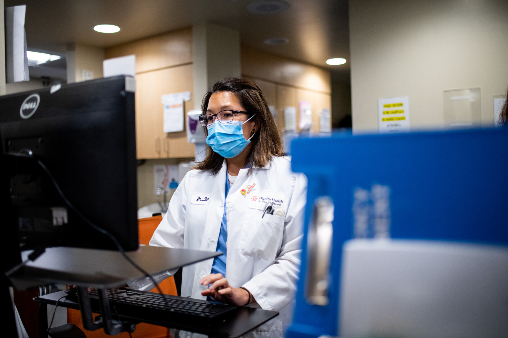 A woman with a white lab coat and face mask standing at a computer.