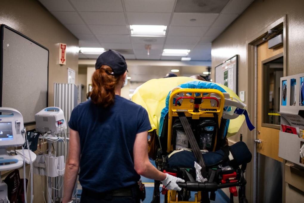 A female EMT pushes a patient on a gurney through an emergency room.