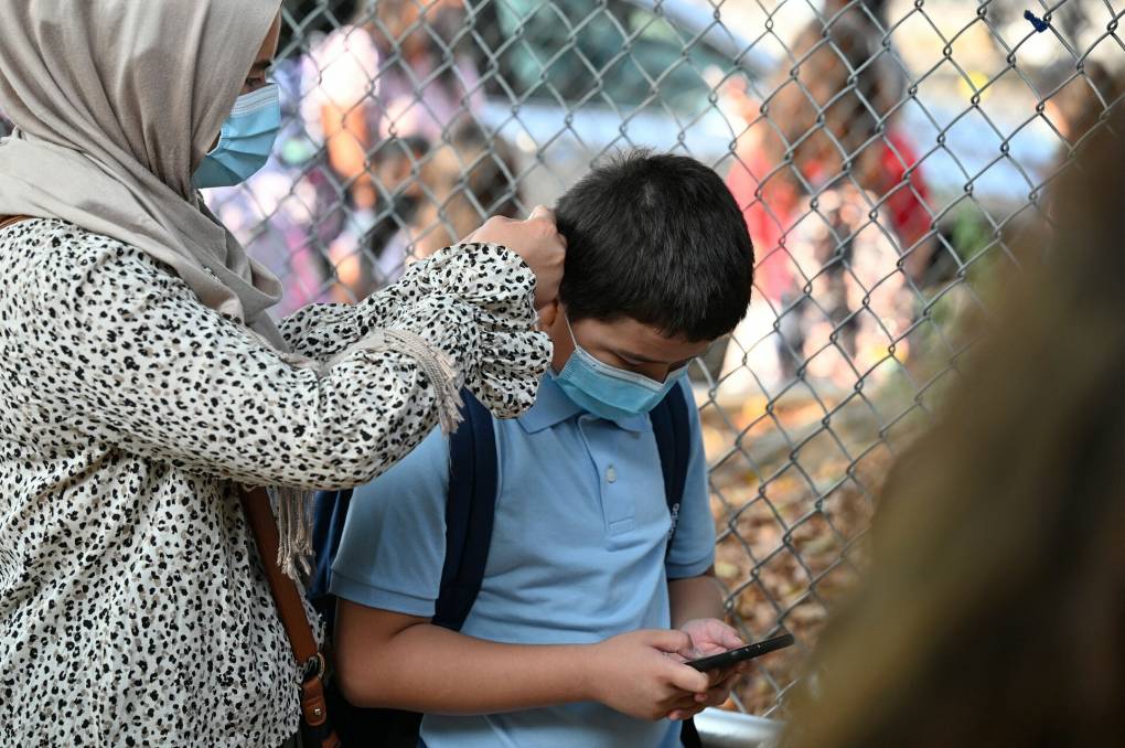 A parent wearing a beige-colored hijab and patterned shirt adjusts her son's blue face covering outside the gates to his school.