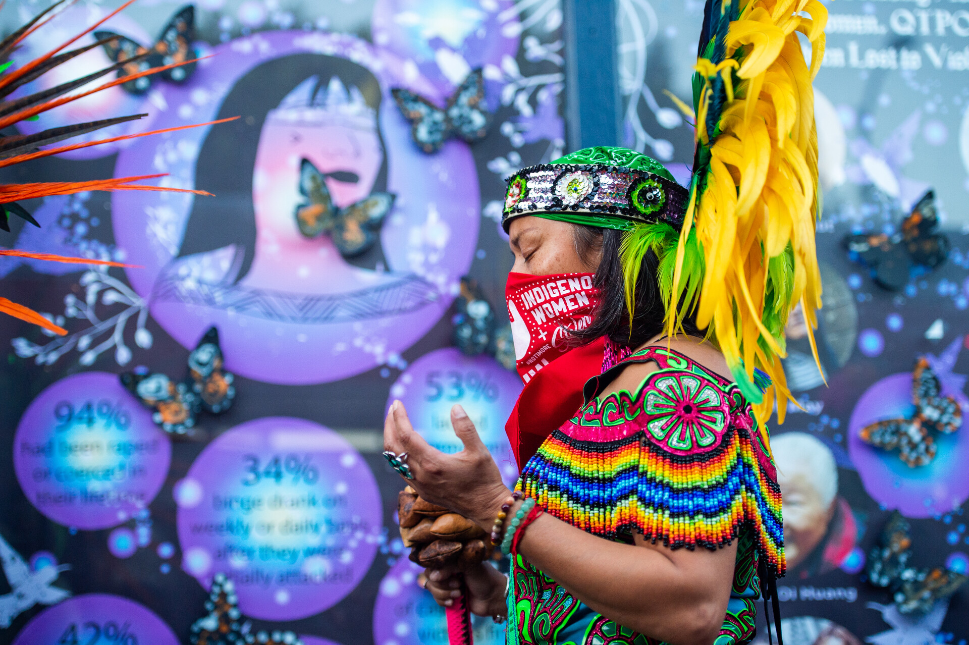 A person wearing traditional Aztec dance regalia lifts their hands up in prayer in front of a poster that shares some of the statistics on violence committed against women and queer and trans people of color.