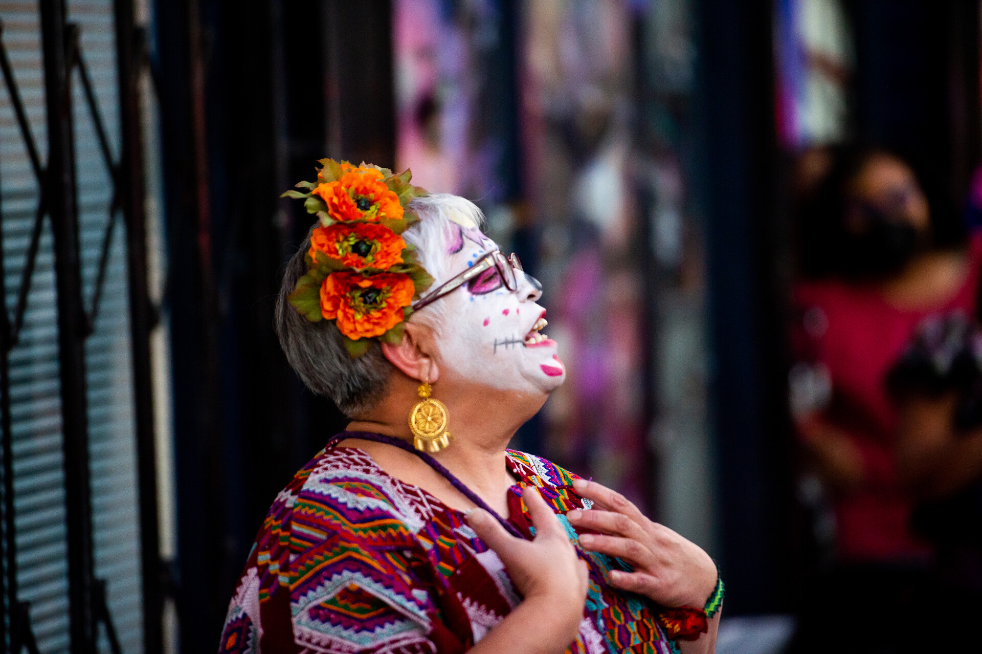 A woman wearing face paint and cemaspúchil in her hair sings to a crowd on the sidewalk.