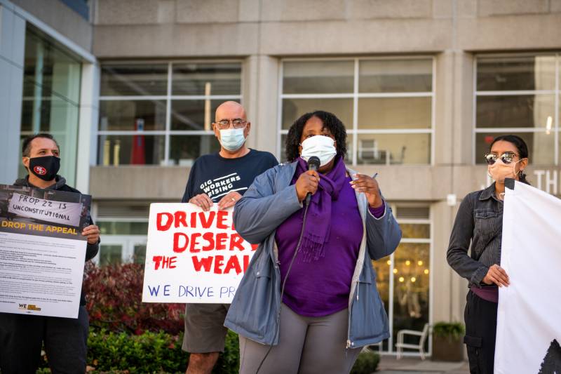 Two men holding signs and two women, one wearing purple and holding a microphone and one woman holding a sign are standing in front of a building.