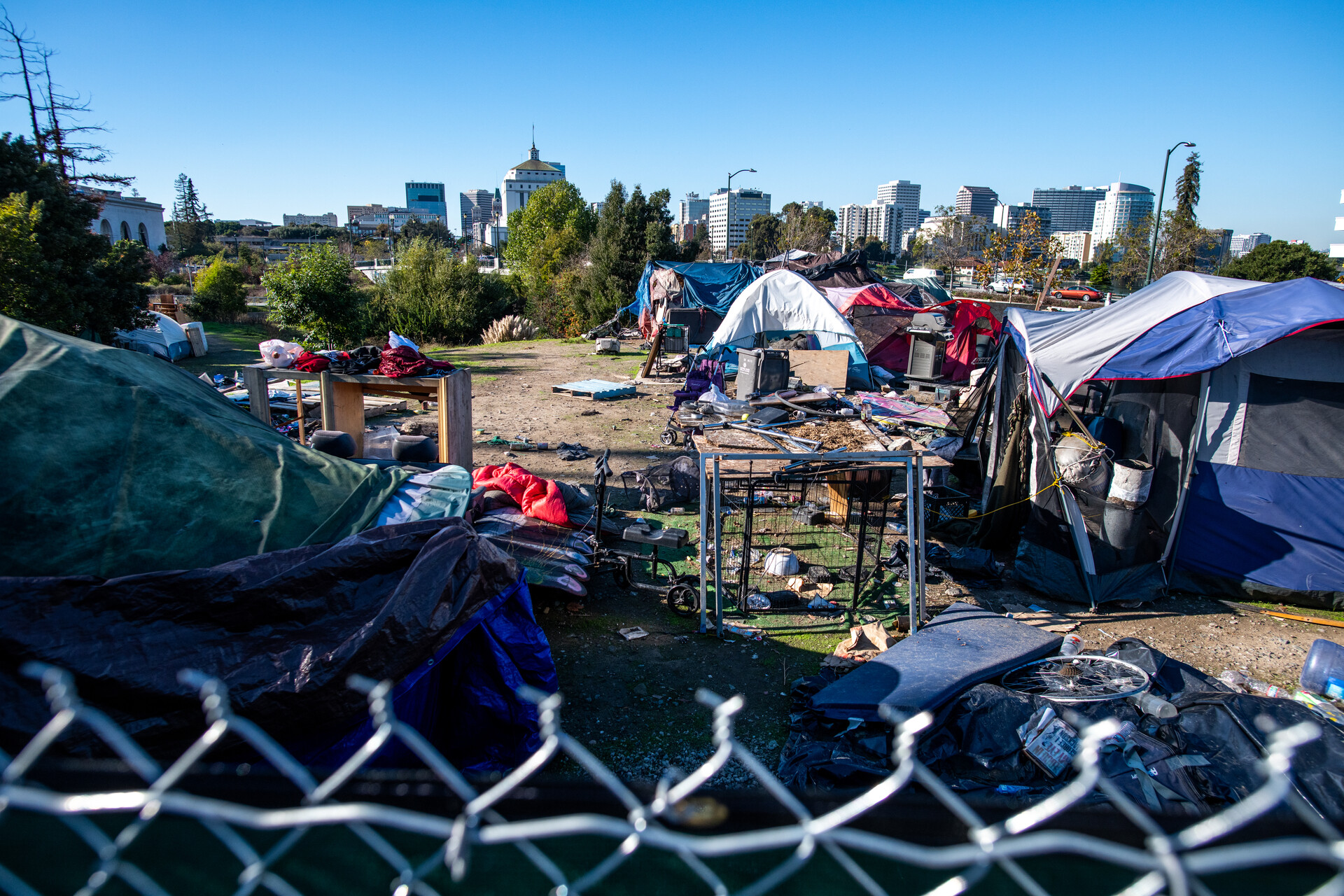 A tent encampment with miscellaneous items all over the ground. In the far background behind a row of trees is Oakland's skyline.