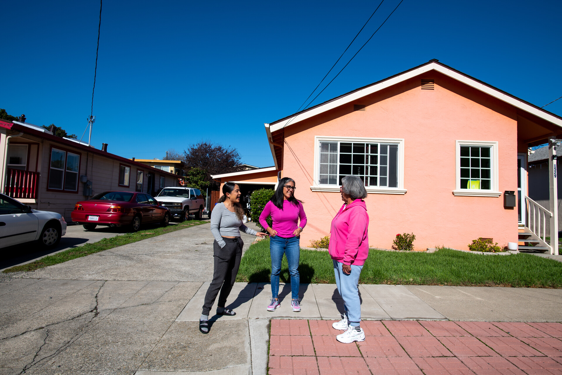 Three women stand by each other having a conversation on the sidewalk in front of a pink house.
