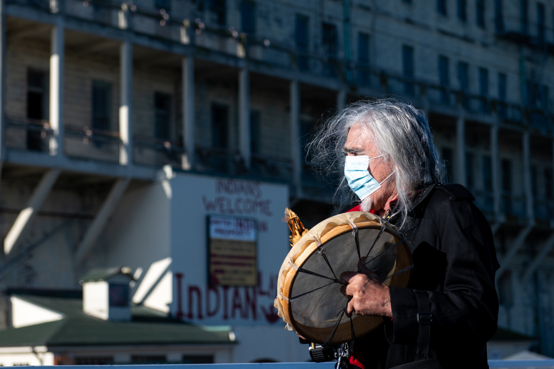 A man with long gray hair bangs a ceremonial drum outside a former prison building on Alcatraz Island.