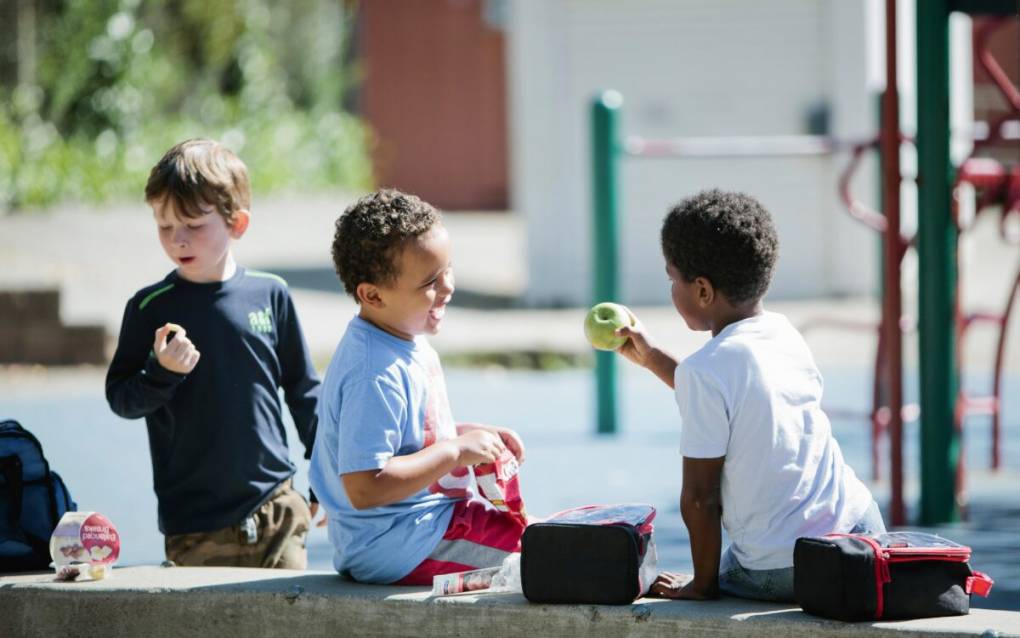 Three children at a playground. One is in the background, and one holds up a green apple at the third child, who holds a red lunch box.