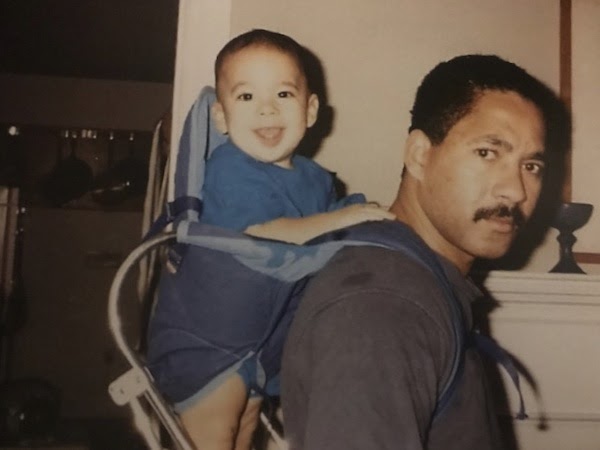 A father who "Black Cuban" sports close cropped hair and a mustache and a gray T-shirt, profiled from the side and looking at the camera. On his back is a smiling baby with lighter skin in a baby backpack, wearing a blue T-shirt. 