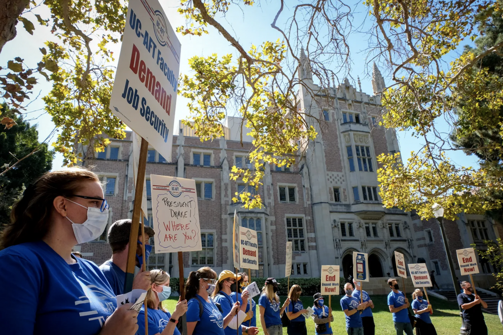 15 people are outside standing on grass wearing blue shirts. 12 people are holding signs. One of the signs reads "UC-AFT Faculty require job security."