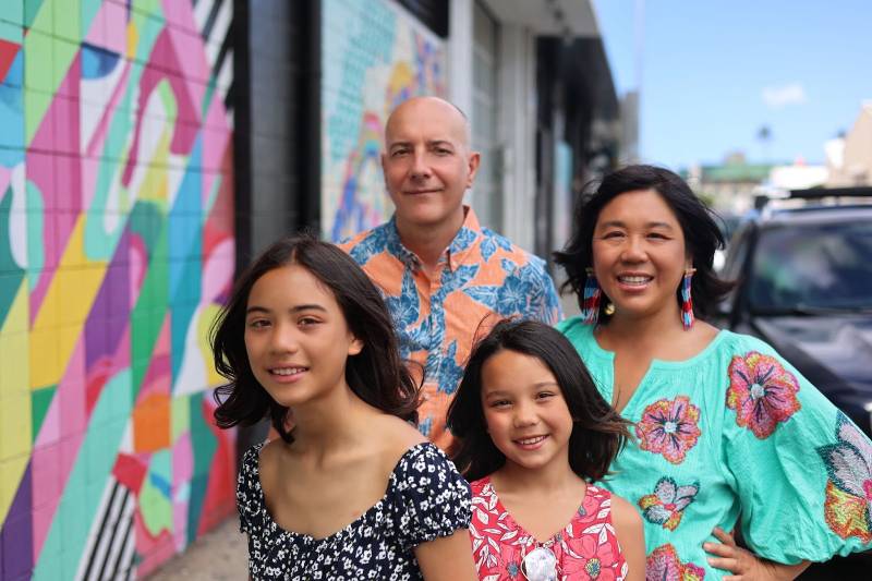 A multiracial family, all decked out in mulitcolored Hawaiian style shirts. They're standing on a street with a similarly multi-colored mural behind them. The Argentinian father is bald, standing in the back on the left, with a bit of a serious look, the mother on the right, also in the back, is of multiracial Chinese and Latina descent, smiling with long earrings and shoulder-length black hair. Their two daughters stand in front of them, each sporting smiles and shoulder-length dark hair. 