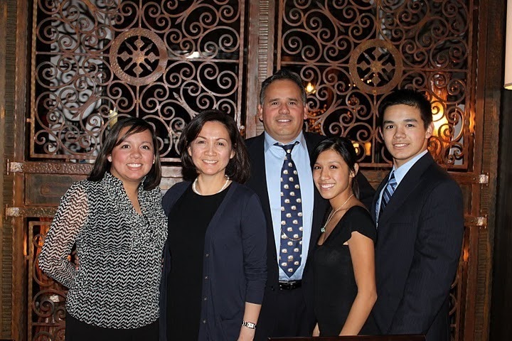 A family of five stand, well-dressed, against an ornate door. They're all smiling. From left to right: a woman in a black and white patterned top with her arms behind her back, an older woman (the mom) in a dark blue blouse with a black undershirt and white necklace, a taller man (the dad) in a light blue dress shirt and navy blue patterned tie (his hair has specks of gray), a shorter woman in a simple black dress and a necklace with a pendant, a young man in a suit and tie similar to the older man's. 