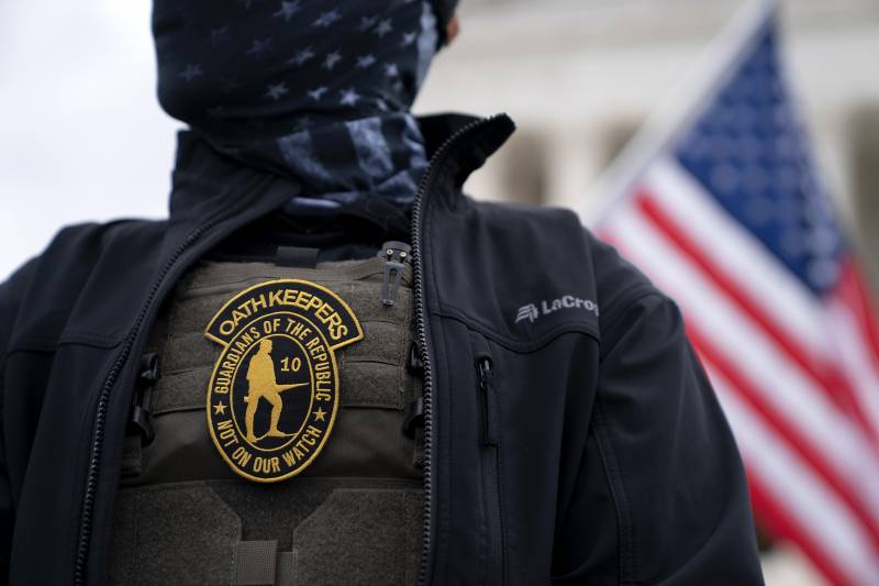 A man whose lower face is completely concealed wears an "Oath Keepers" badge on a bulletproof vest beneath a black windbreaker.