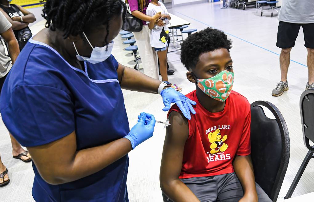 A nurse wearing scrubs and a mask injects a needle into a child's arm who is seated.