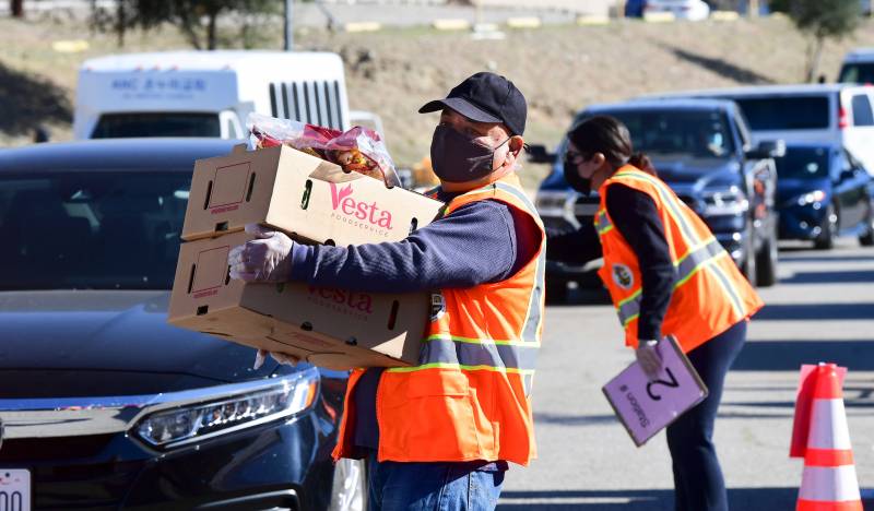 Two people wearing bright orange safety vests and masks. One holds two boxes of produce on his way toward what looks like a line of cars, while the woman behind him holds a clipboard and leans forward to speak into what looks like an open car window.