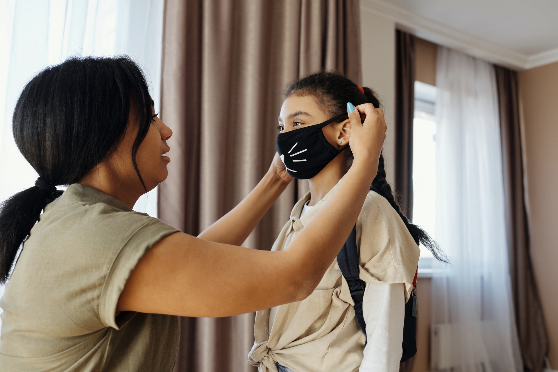 A woman helps a young girl put on a black cloth face mask with cat whiskers.