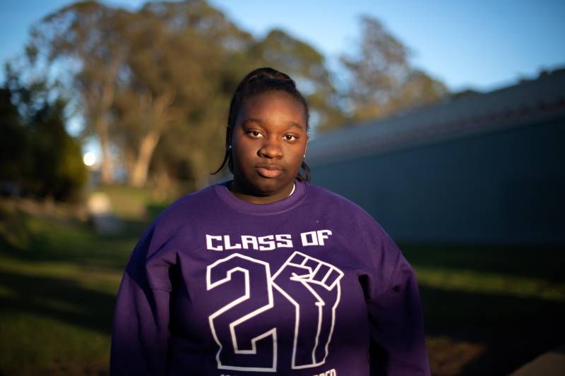A woman stands in the foreground in a purple sweatshirt with the words "Class of 21" with trees and sky in the background.