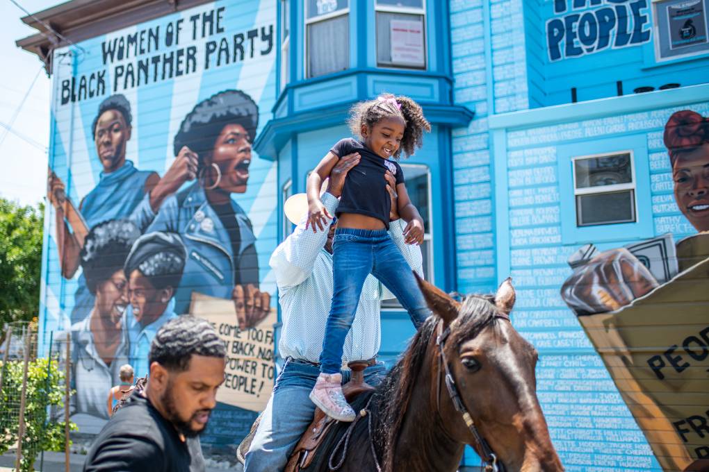 A child smiles as she is lifted onto a horse by a man in a cowboy hat in front of a blue mural that says, "Women of the Black Panther Party."