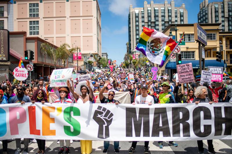 A colorful scene with a large banner in the foreground and many people in the background waving rainbow flags and signs.
