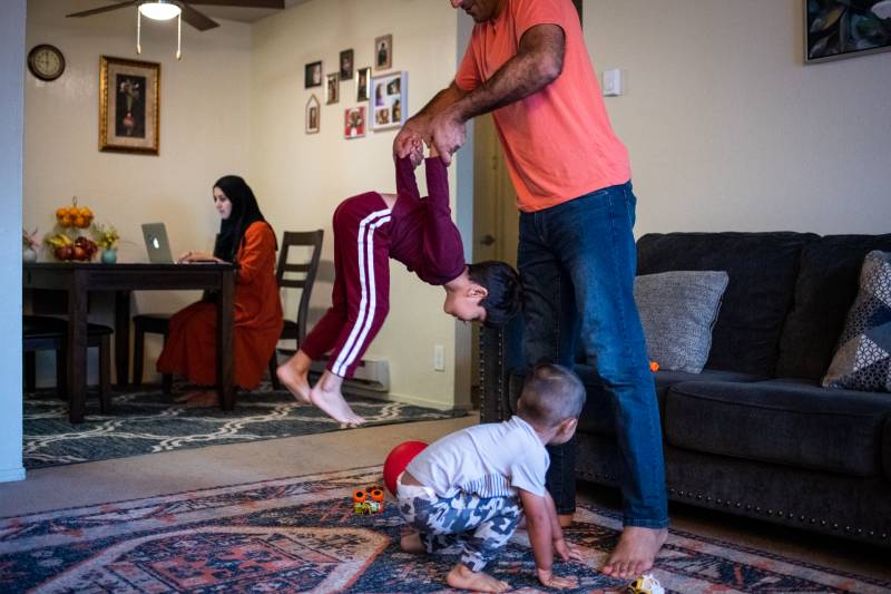 A man holds a child by the hands as the child flips upside down inside of a house. Another child plays nearby on the floor while a woman works on a computer at a kitchen table in the background.