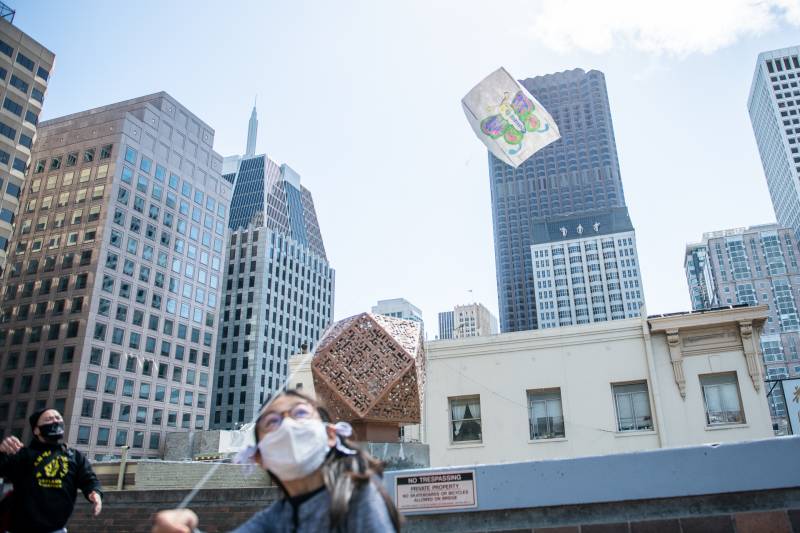 A girl with a mask in the bottom left of the frame flies a kite with the background of buildings in San Francisco 