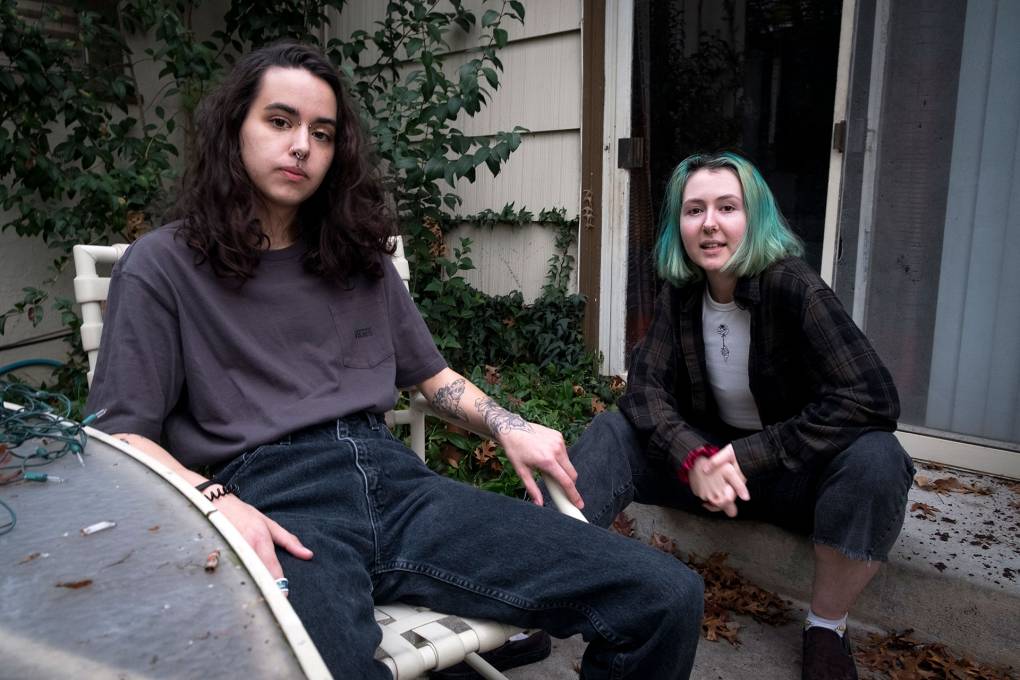 two young students, one with long dark hair, the other with short dyed blue hair, look pensively at the camera on their patio