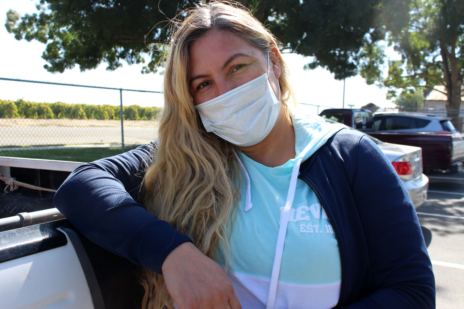 A light-complected woman with long blond hair looks frankly at the camera, her right elbow propped on top of a car, her head tipped to her right. She wears a white mask that covers her face from nose to chin, a teal and white sweatshirt, and a dark blue jacket. Beyond her and beneath a leafy tree, a couple other parked cars are visible, and beyond a chain link fence lies a field with brown dirt and bright green leafy crops.