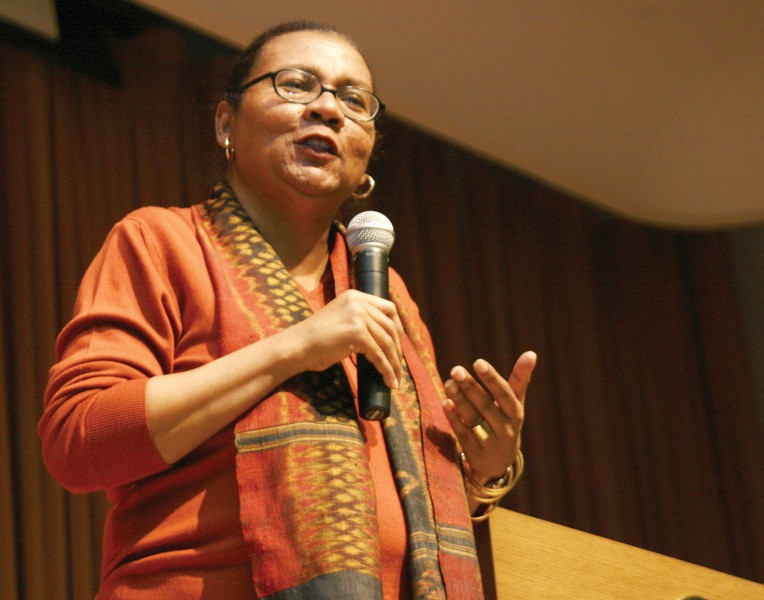 bell hooks stands next to a podium wearing an orange long-sleeve shirt and scarf speaking with a mic in her right hand.