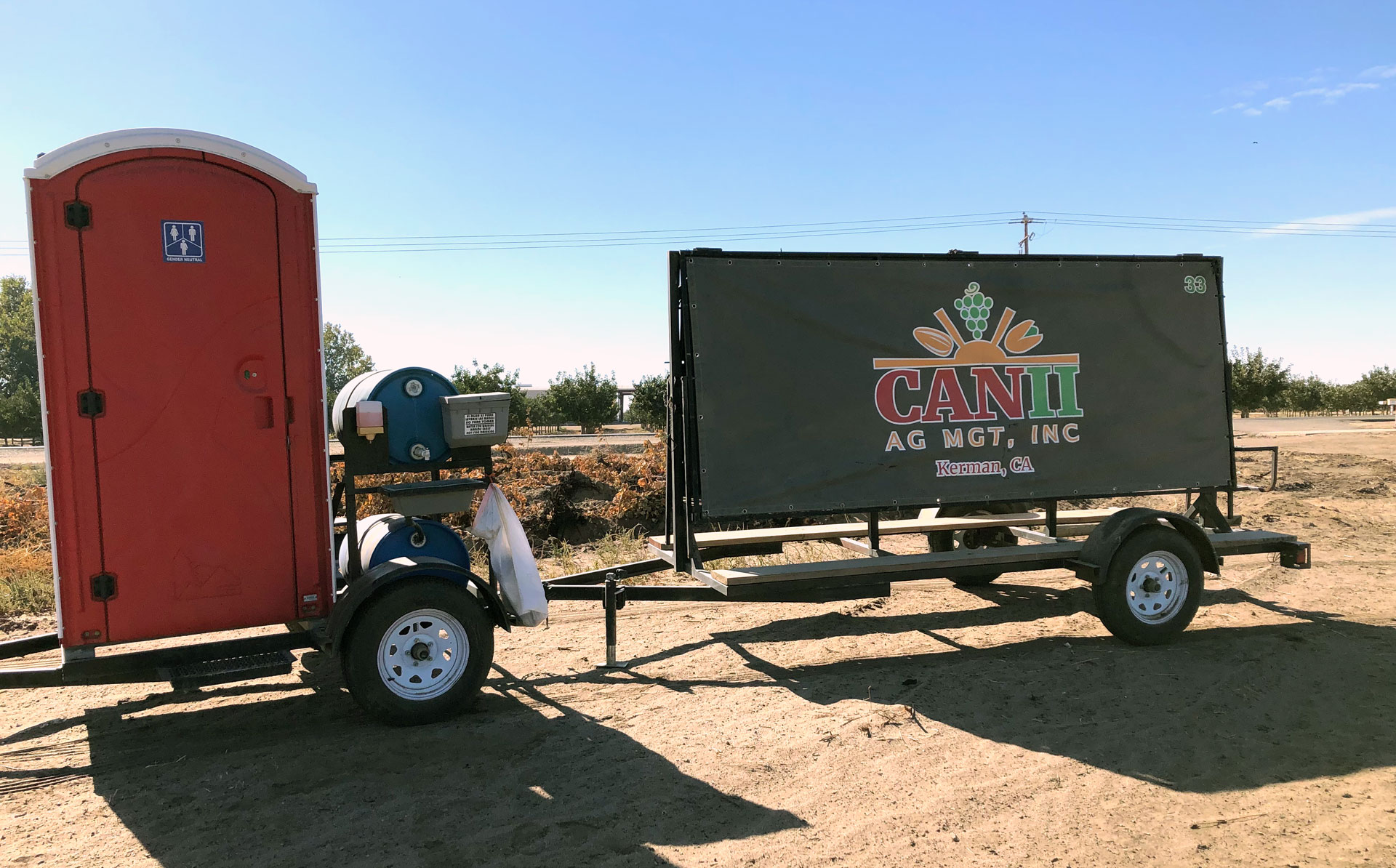 Backlit by bright sun on a four-wheeled extended trailer are a red portable toilet with a hand-washing station on the left and an olive-colored company sign nearly the height of the toilet and three times as long on the right. The trailer sits on a dirt road beside a field.