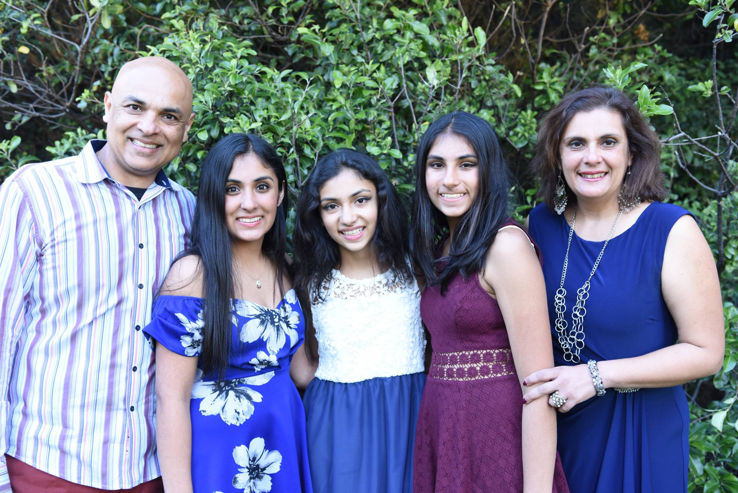 A family snapshot taken outdoors features a smiling father, mother and three teenage girls.