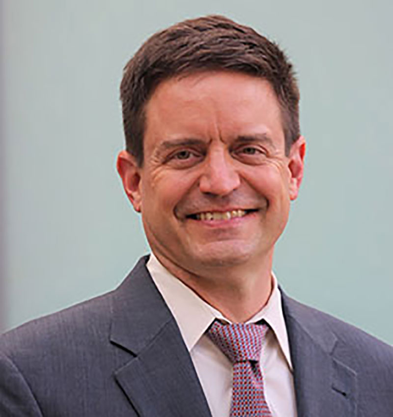 A professional head shot of a smiling, middle-aged White man with medium-short brown hair, a gray suit jacket, pink shirt, and darker pink tie.
