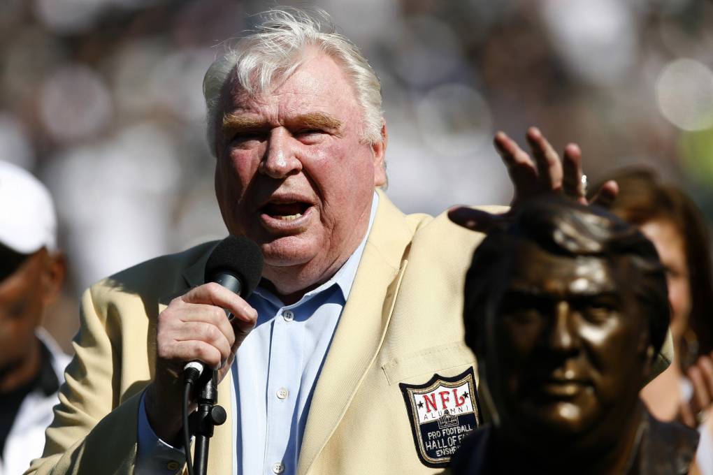 John Madden stands outside and speaks through a microphone. He stands next to a bust of his face made of bronze.