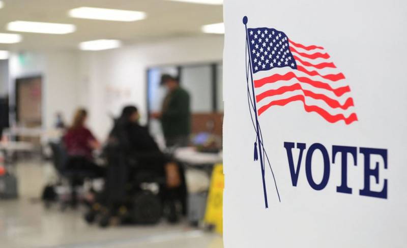 A sign on a booth with an American flag has the word "Vote" written on the side. There are two people sitting and one person standing in the blurred background.