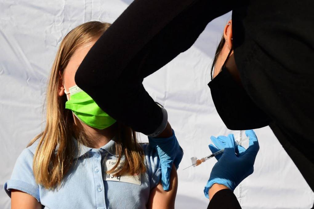 A child wearing a green mask and blue shirt receives a needle in her arm from an adult wearing surgical gloves and a syringe.