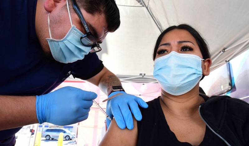 A man wearing glasses, a surgical mask and gloves injects a syringe into the arm of a woman wearing a black shirt and surgical mask.