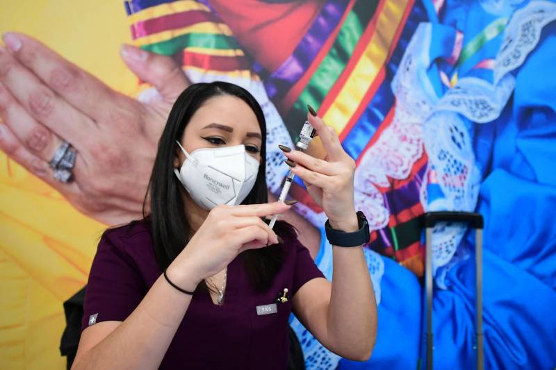 Eloisa Flores wears a face mask as she fills a syringe with a vaccine dose. She works outside and behind her is a mural.